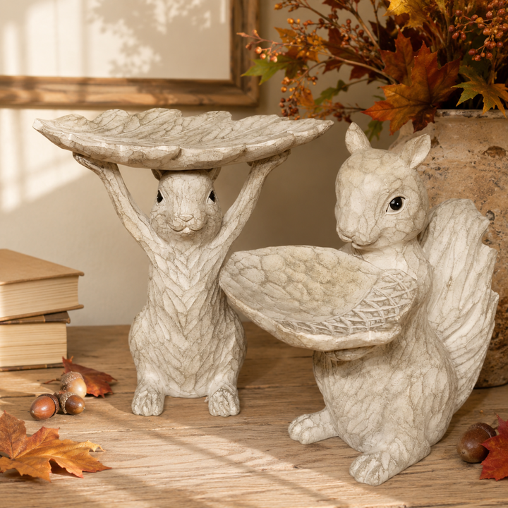 Two faux stone squirrel garden figurines in a warm neutral setting. One squirrel holds a leaf-shaped tray overhead, and the other sits beside it holding a matching leaf tray.