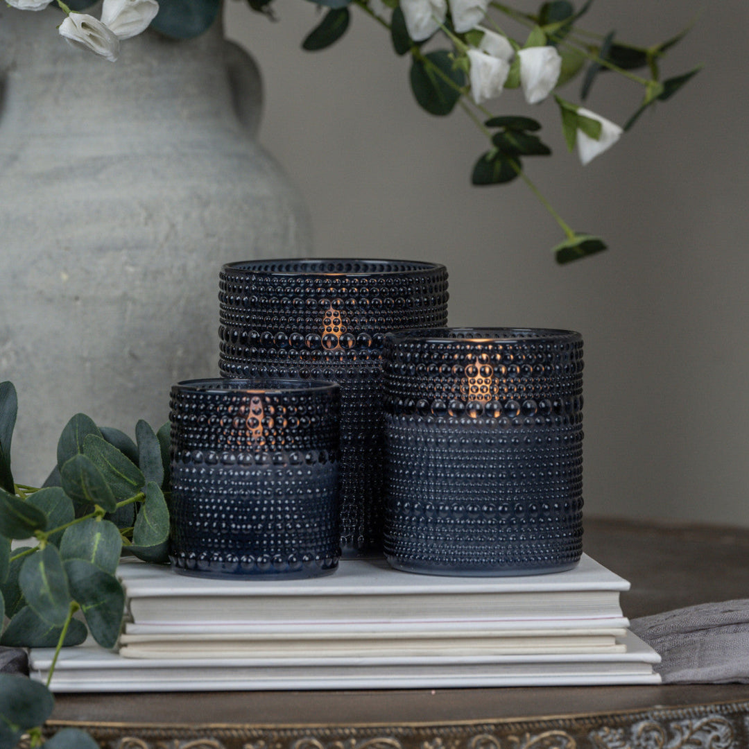 Set of dark blue textured candle holders on a stack of books with a vase and greenery in the background.