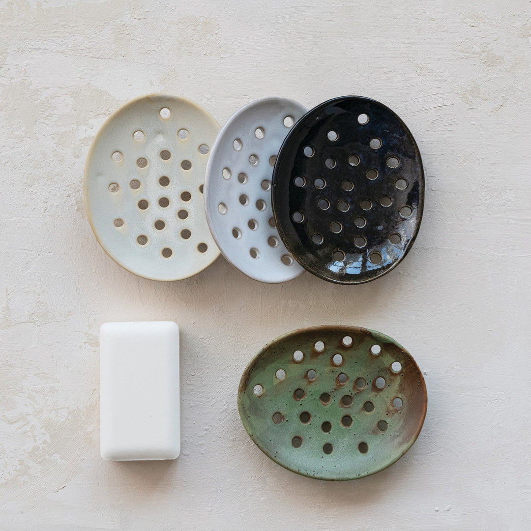 Five ceramic soap dishes in different colors on a light surface with a bar of soap.