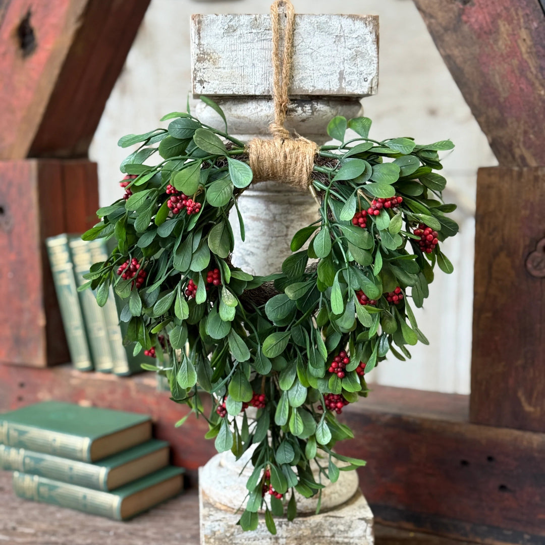 Decorative wreath with green leaves and red berries on a wooden stand against a rustic background.