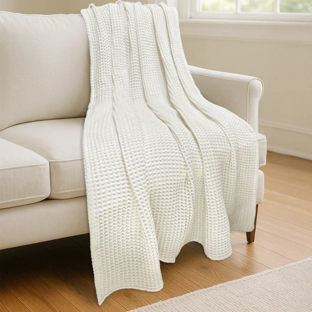 A cream-colored waffle-knit throw blanket draped over a light beige sofa in a bright, modern living room with light wood floors.