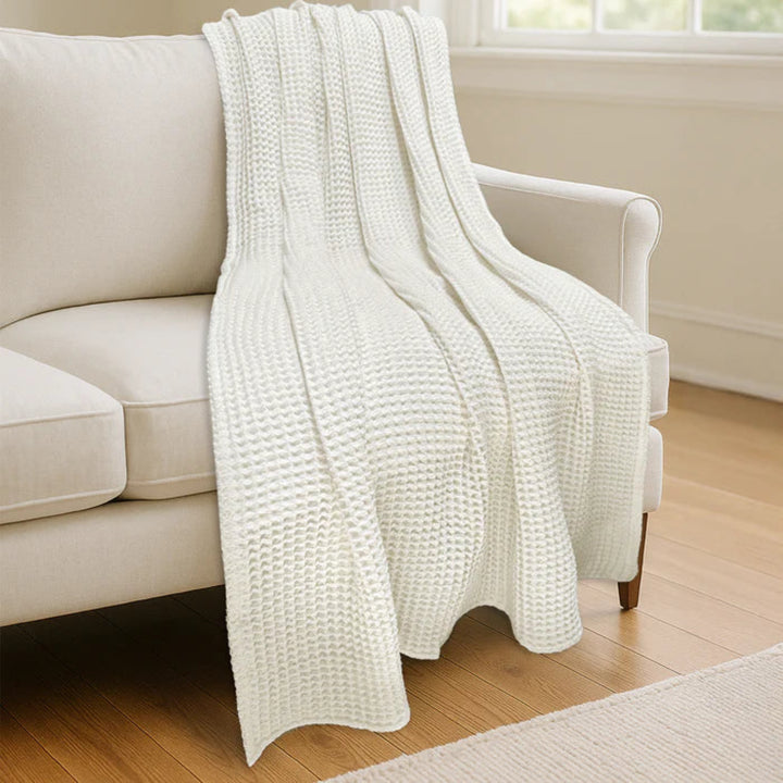 A cream-colored waffle-knit throw blanket draped over a light beige sofa in a bright, modern living room with light wood floors.