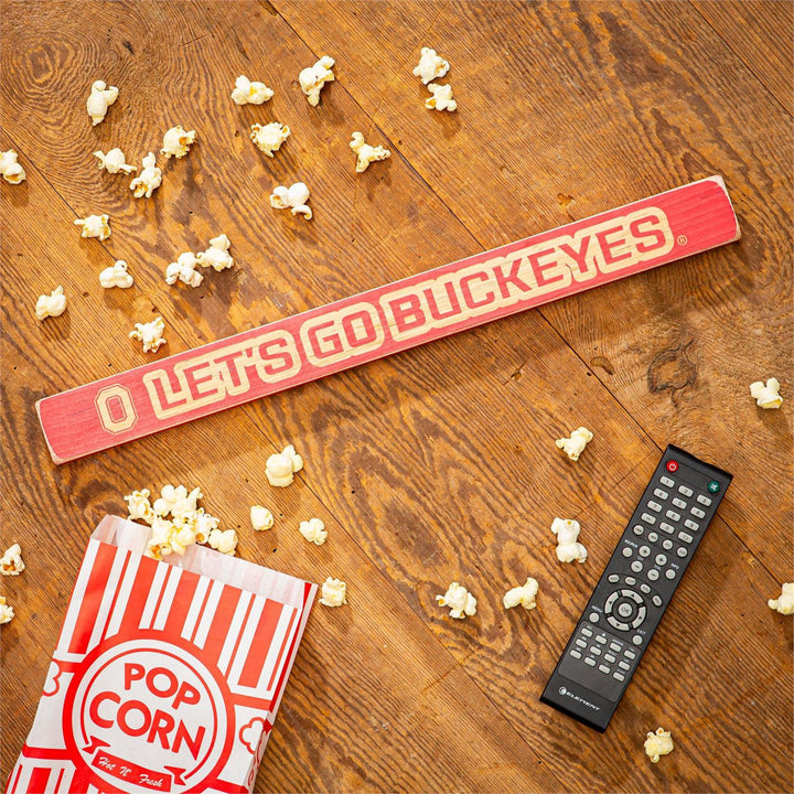 Wooden sign with 'O Let's Go Buckeyes' on a wooden floor with popcorn and a remote control.