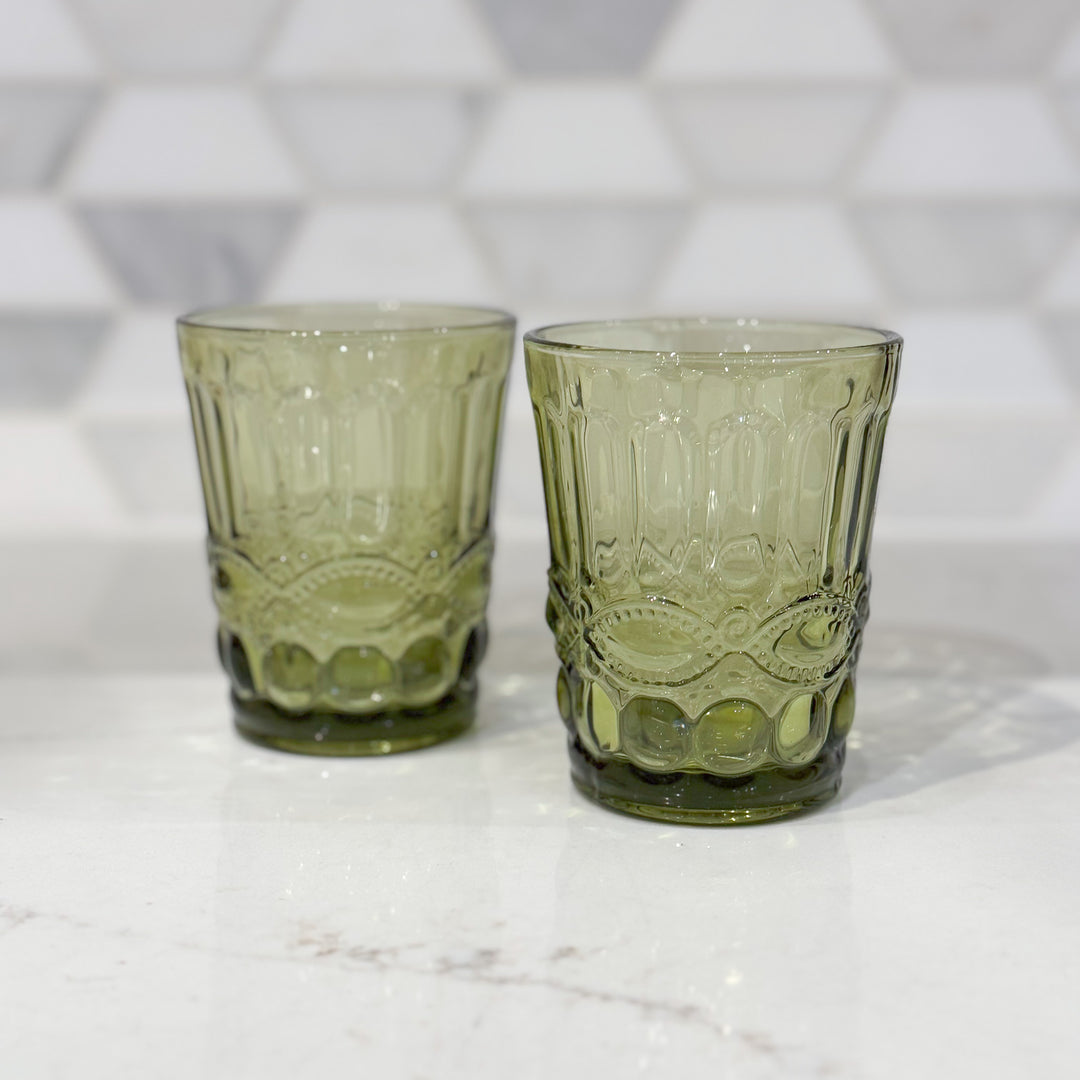 Two embossed olive green glass drinking glasses with a geometric pattern, placed on a marble surface.