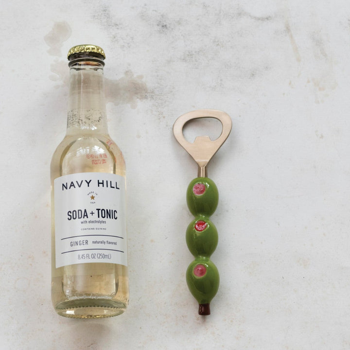 Bottle of Navy Hill Soda & Tonic with a bottle opener and olives on a marble surface