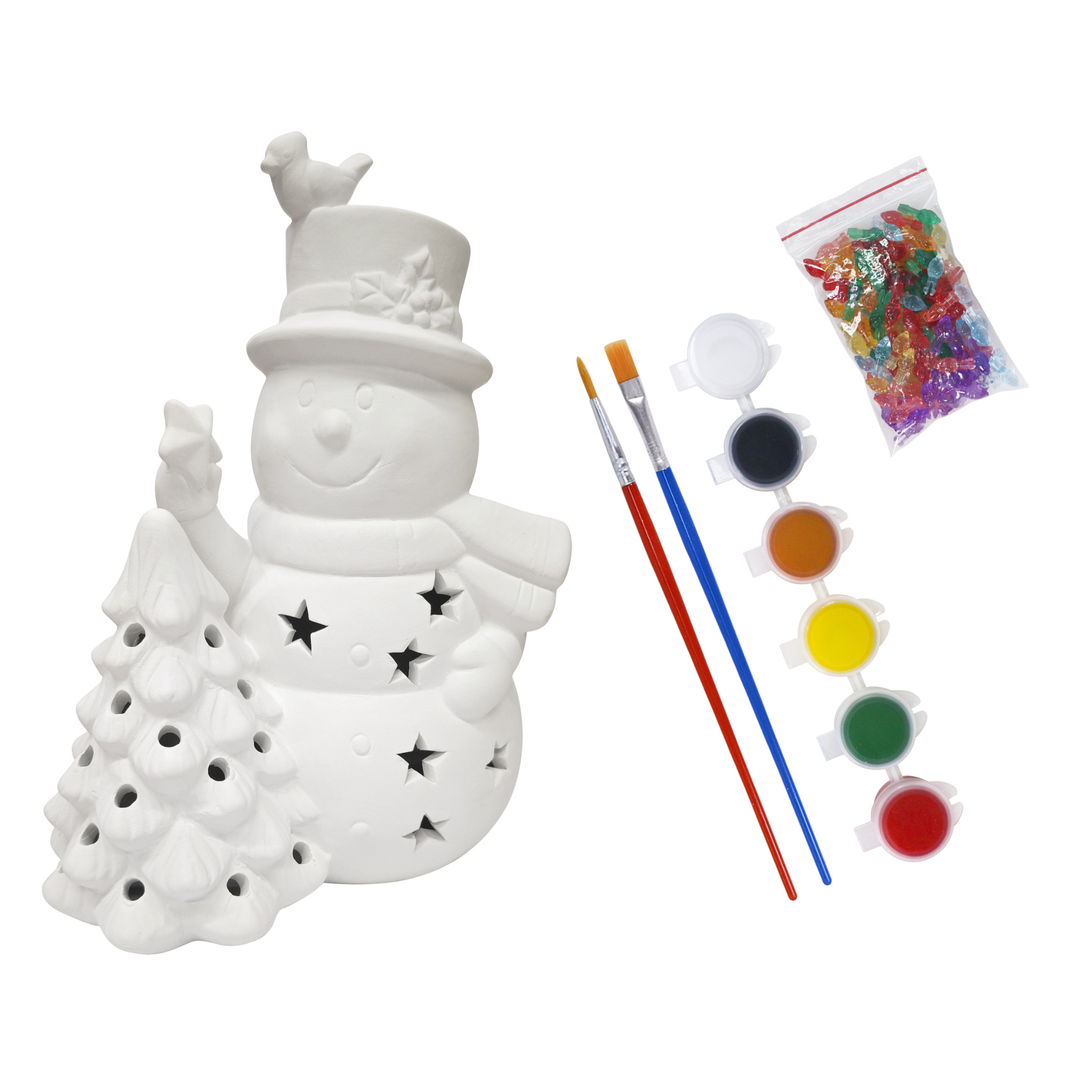 White ceramic snowman and Christmas tree craft kit with star cutouts, shown with paintbrushes, small paint pots, and a bag of colorful glitter on a white background.
