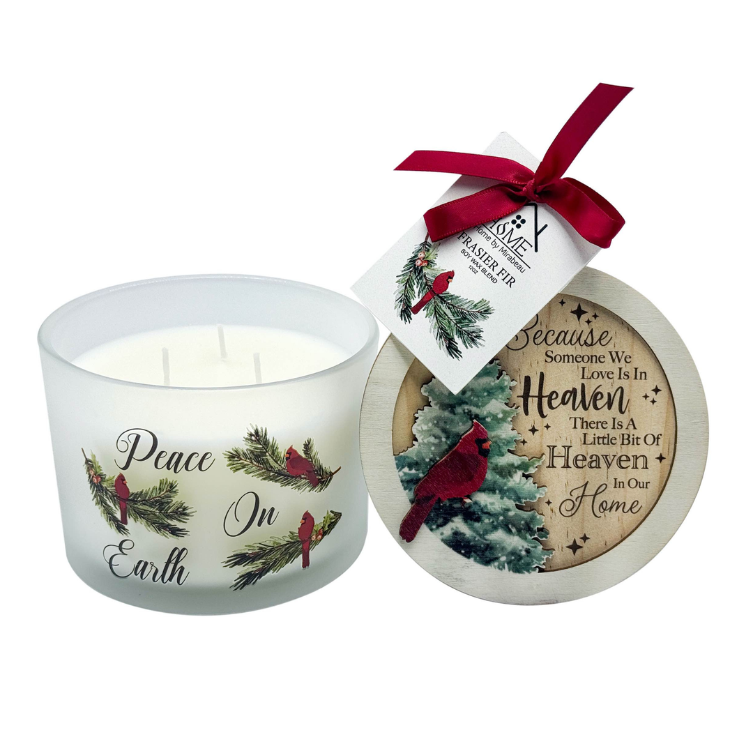 A holiday-themed scented candle in a frosted glass jar with 'Peace In Earth' text and cardinal bird illustrations, shown next to its round lid. The lid features a printed holiday design with a red cardinal, pine branches, and the quote 'Because Someone We Love Is In Heaven There Is A Little Bit Of Heaven In Our Home.