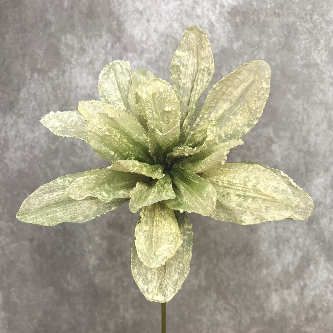A single artificial flower stem with wide, textured pale green and yellow-green petals on a dark stem, set against a mottled gray studio background.