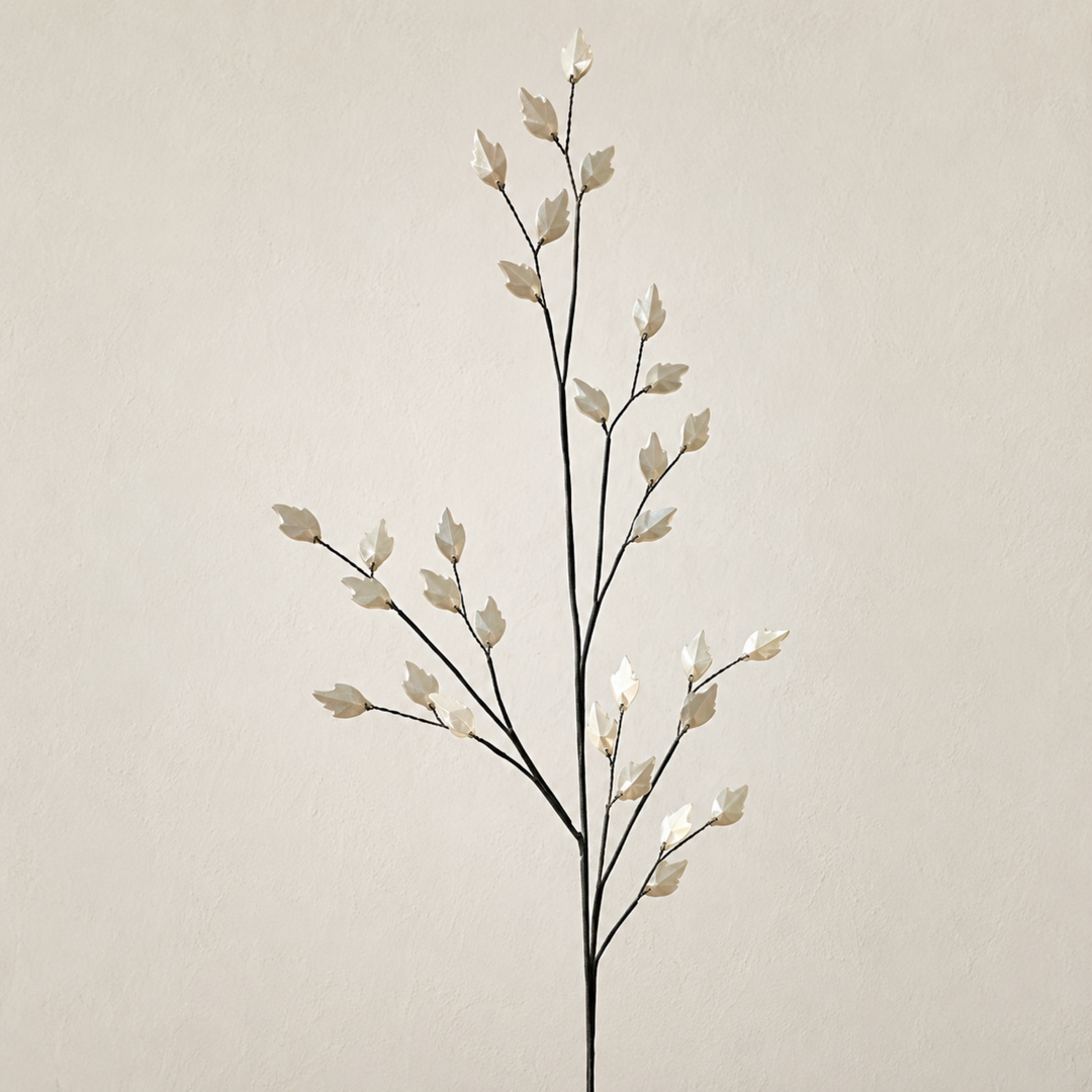 A decorative branch-style wall decor piece with slender dark stems and small glossy off-white leaf-like tips, shown against a plain warm beige wall.
