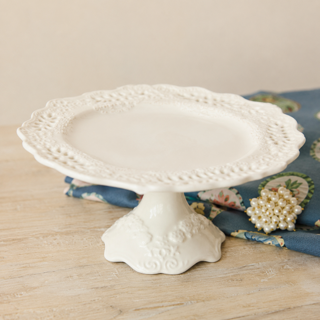 White decorative cake stand on a wooden surface with a floral patterned fabric in the background.