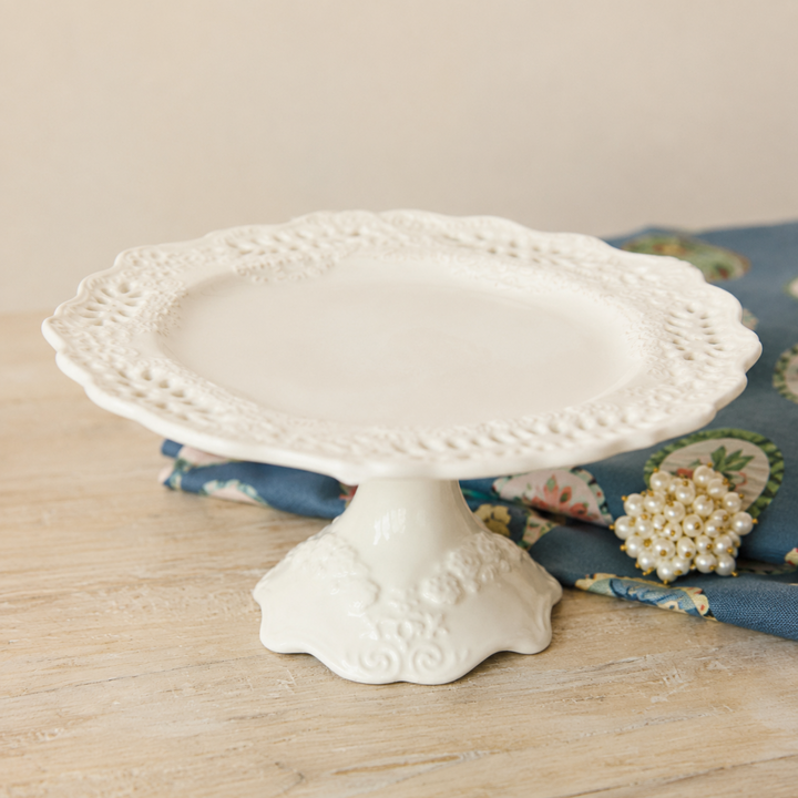 White decorative cake stand on a wooden surface with a floral patterned fabric in the background.