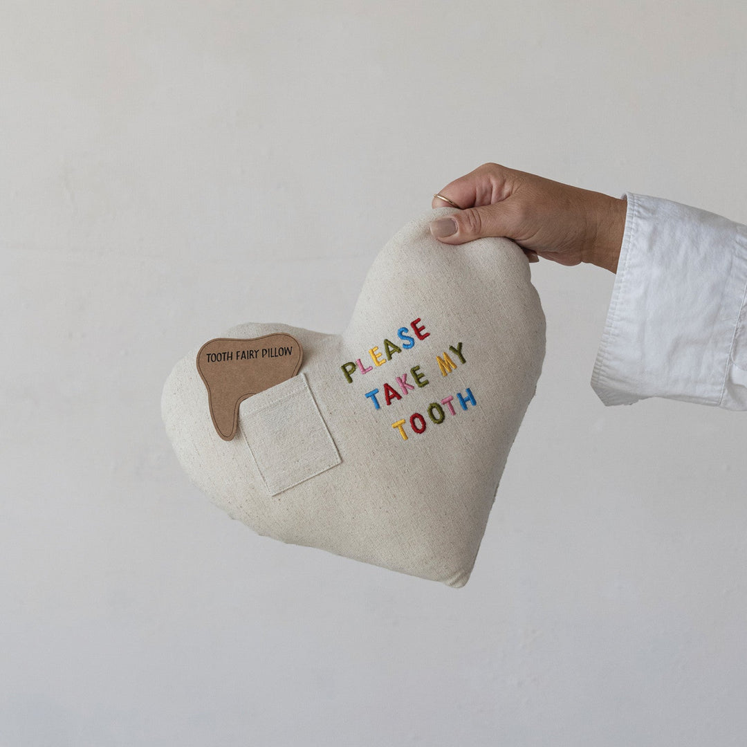 Heart-shaped pillow with 'Please Take My Tooth' text held by a hand against a plain background