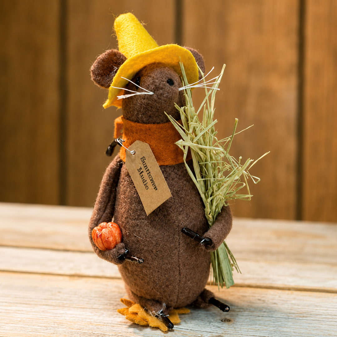A small brown felt mouse figurine dressed as a pumpkin, wearing a yellow hat, orange scarf, and holding a small orange pumpkin and a bundle of hay, standing on a wooden surface against a wooden plank background.