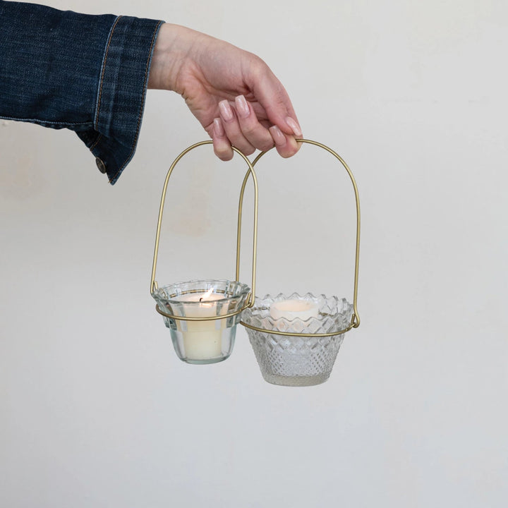 Hand holding a gold metal and glass candle holder with two candles inside on a plain background