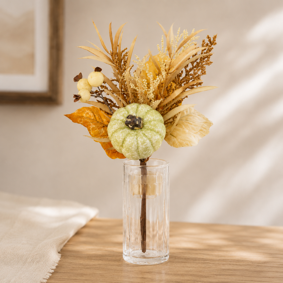 An autumn-themed artificial floral arrangement in a clear ribbed glass vase, featuring a small light green pumpkin, dried yellow and brown grasses, and a single orange leaf, displayed on a wooden tabletop.