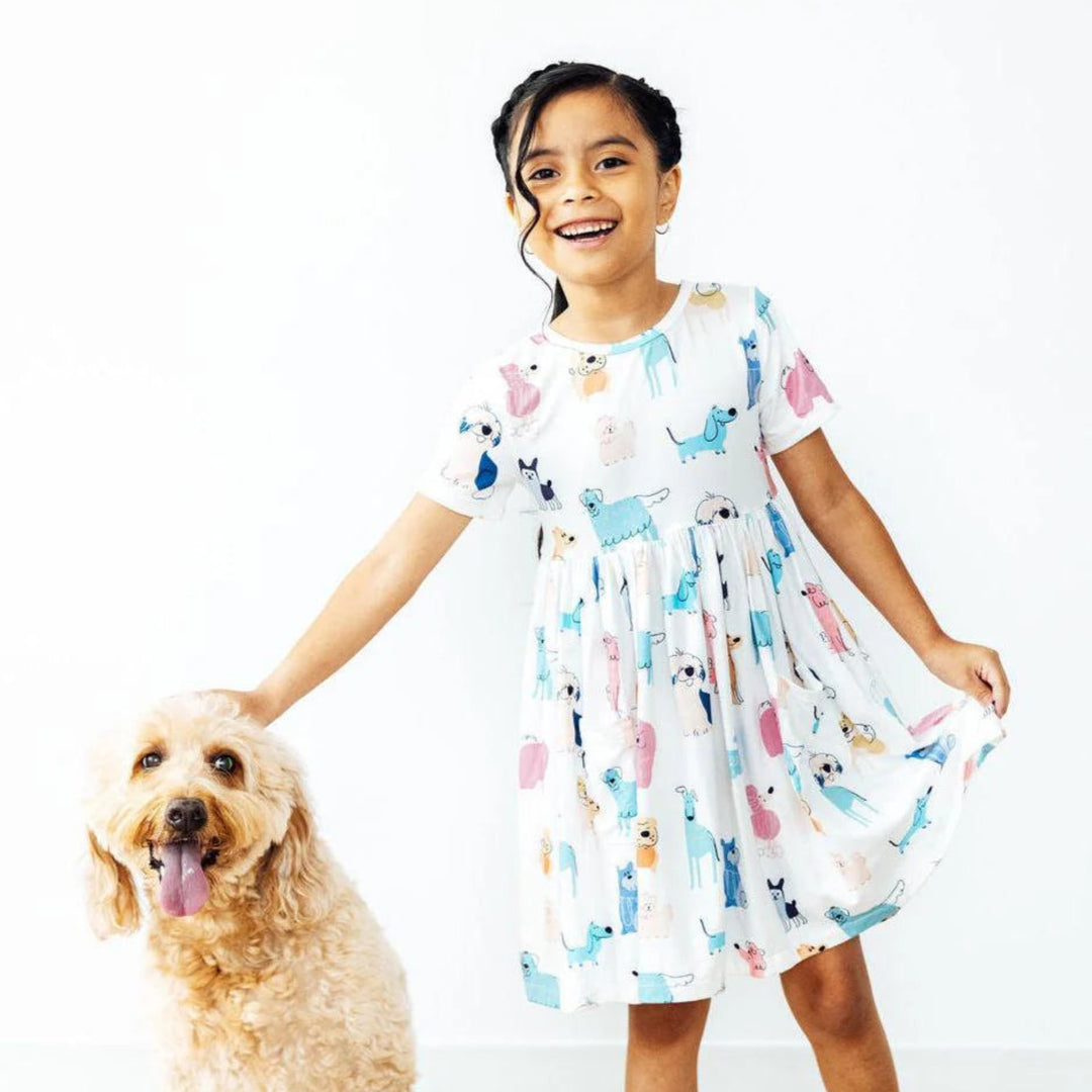 Young girl in a patterned dress standing next to a dog on a wooden floor.