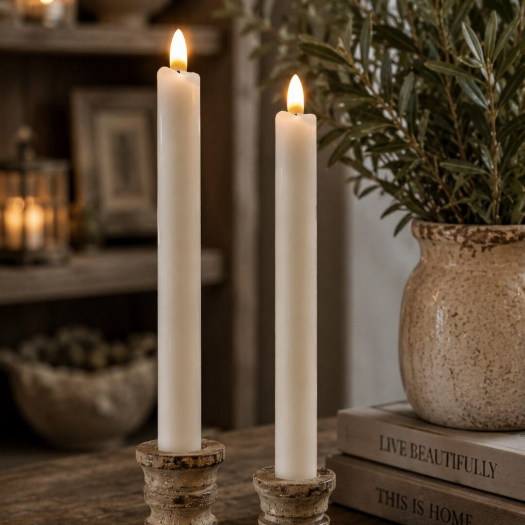 Two lit white taper candles in distressed rustic metal candlesticks on a wooden table, with a potted olive branch and stacked books in a softly blurred home interior background.