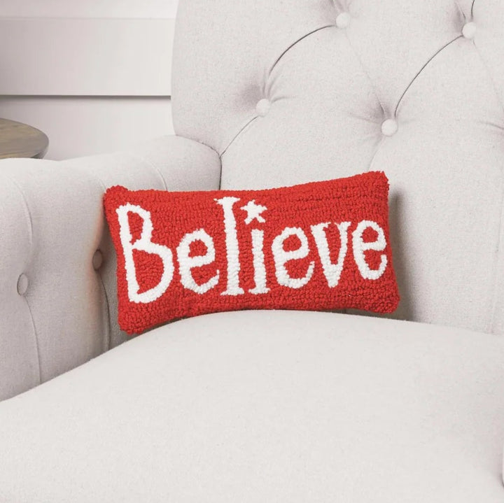 A red rectangular throw pillow with the word 'Believe' in white embroidered letters sits on a light beige tufted armchair in a styled room setting next to a wooden side table with books and a potted plant.