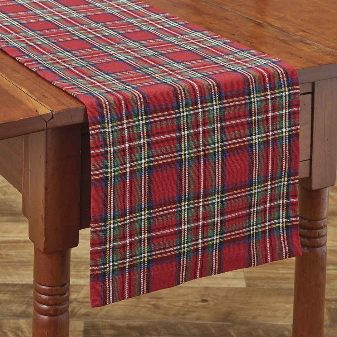A red plaid table runner draped over a wooden dining table, featuring intersecting stripes of green, blue, white, and yellow.