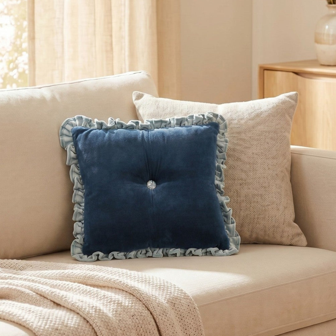 A navy blue velvet square throw pillow with a ruffled trim and a small central button, styled on a beige sofa in a warm, sunlit living room.