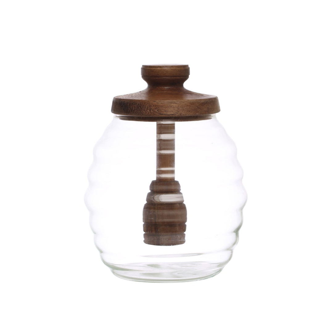 A clear glass jar with a wavy ribbed design, a dark brown wooden lid, and a matching wooden honey dipper inside, set against a plain white background.