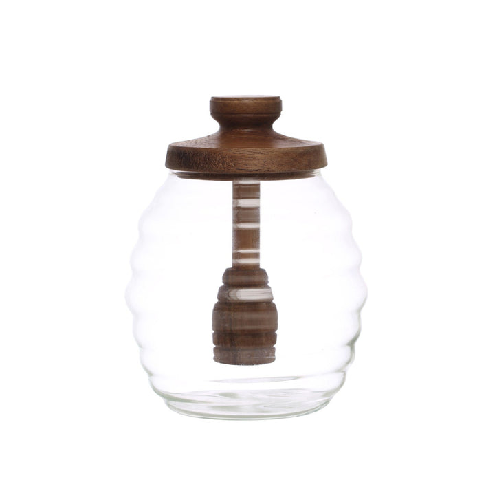 A clear glass jar with a wavy ribbed design, a dark brown wooden lid, and a matching wooden honey dipper inside, set against a plain white background.