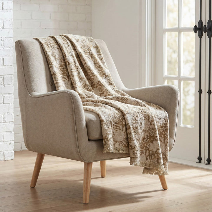 A light beige upholstered armchair with tapered wooden legs, styled in a bright room with a textured cream throw blanket draped over it, set against a white brick wall and French doors.