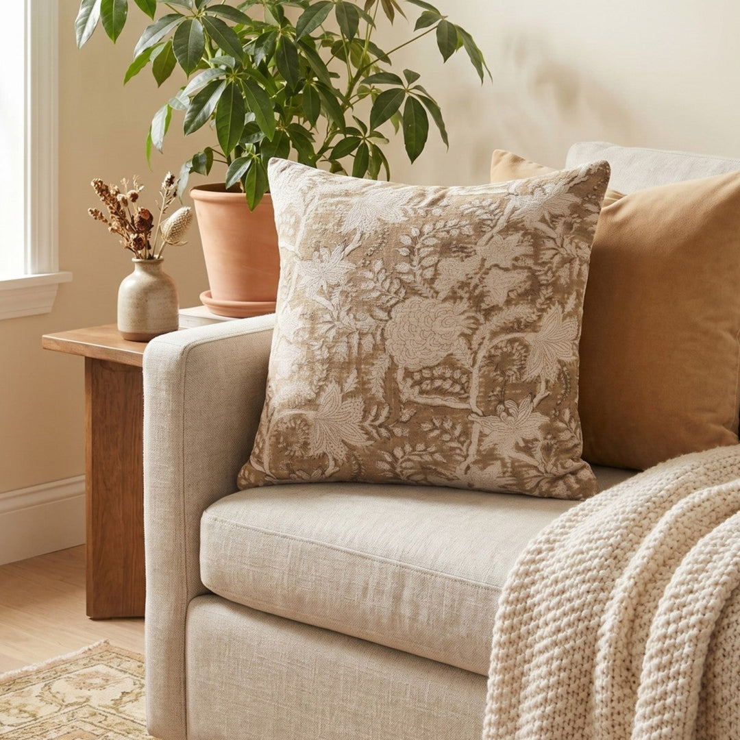 A styled living room scene showing a beige upholstered armchair with a textured cream floral throw pillow and a tan pillow, alongside a cream knit throw blanket, a wooden side table, a potted plant, and a window.