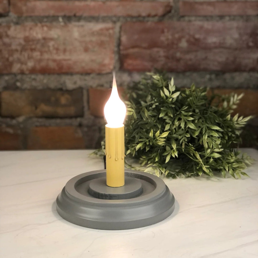 A tabletop candle holder with a gray circular base and a yellow candle-shaped holder supporting a lit flame-shaped bulb, set on a white marble-like surface with a brick wall and greenery in the background.