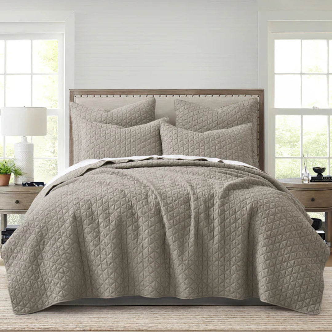 Quilted beige bedding set on a bed in a bedroom with windows and furniture.