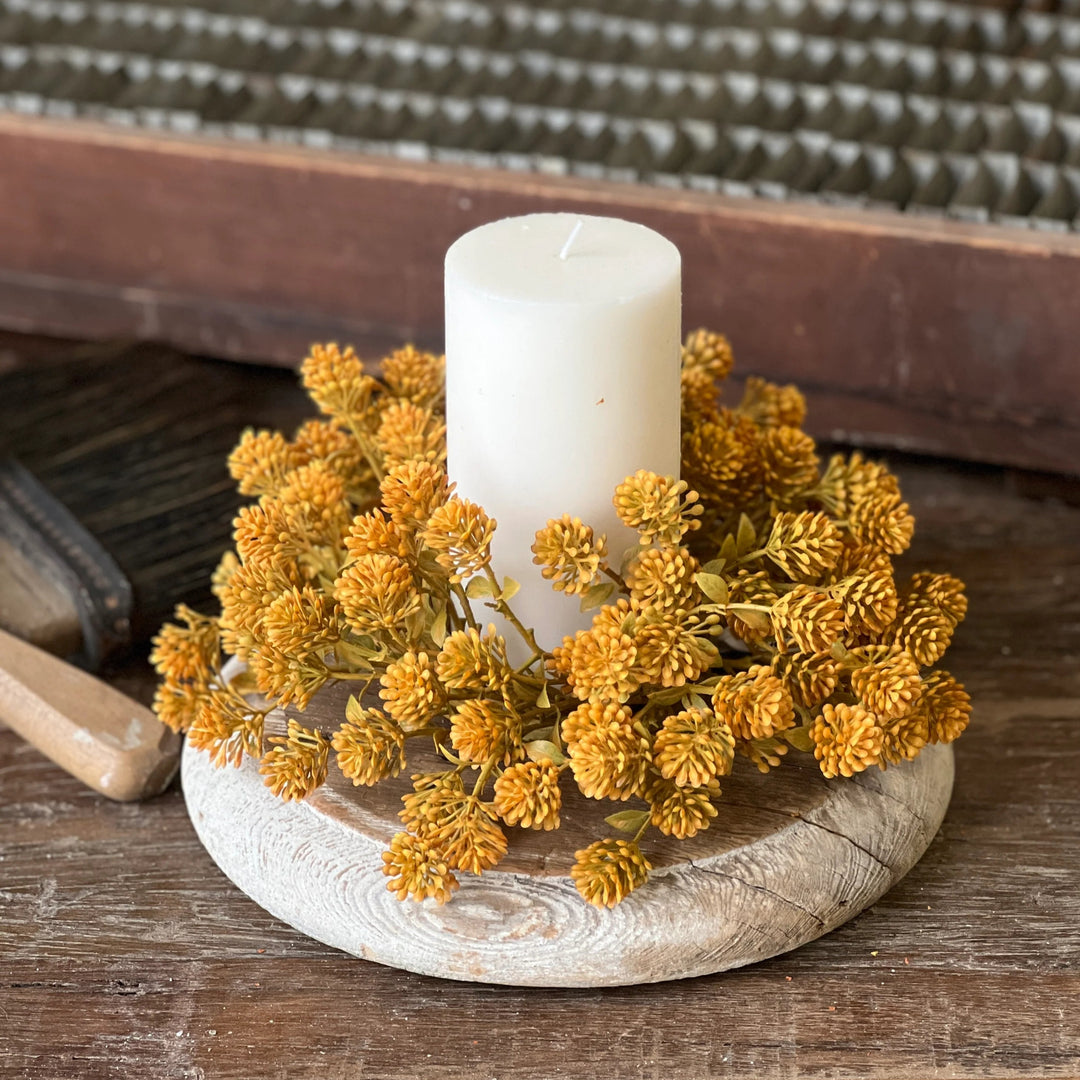 Candle in a decorative holder with yellow flowers on a wooden surface, with a vintage suitcase in the background.