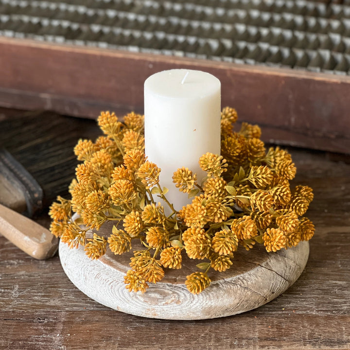 Candle in a decorative holder with yellow flowers on a wooden surface, with a vintage suitcase in the background.