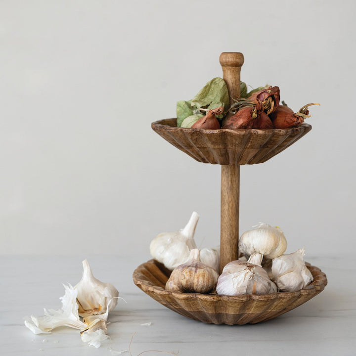 A two-tier wooden serving stand with a central pole and bowl-like tiers, styled with garlic bulbs and dried herbs on a light neutral background.
