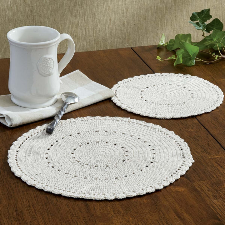 Two white crocheted placemats on a wooden table with a white mug and spoon.