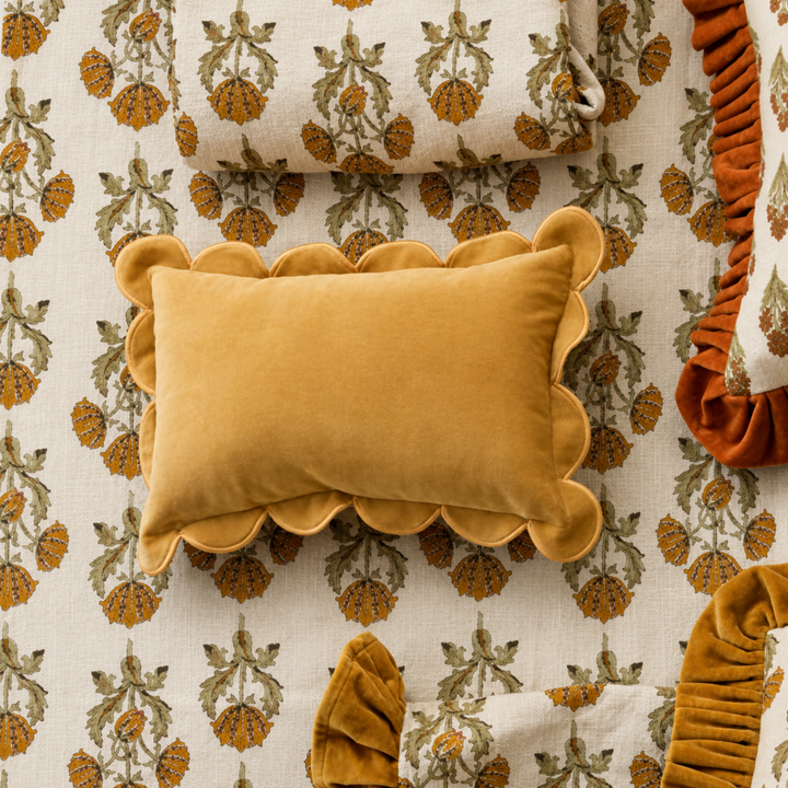 A mustard yellow velvet lumbar pillow with scalloped edges, styled on top of a cream and orange floral-patterned blanket.