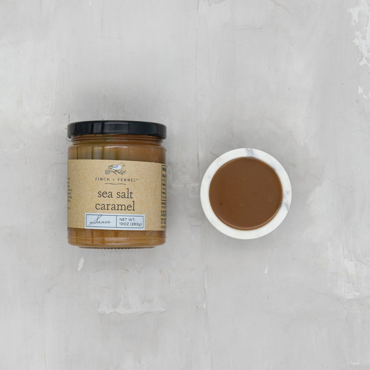 Jar of sea salt caramel sauce with a small bowl of the sauce on a textured white surface