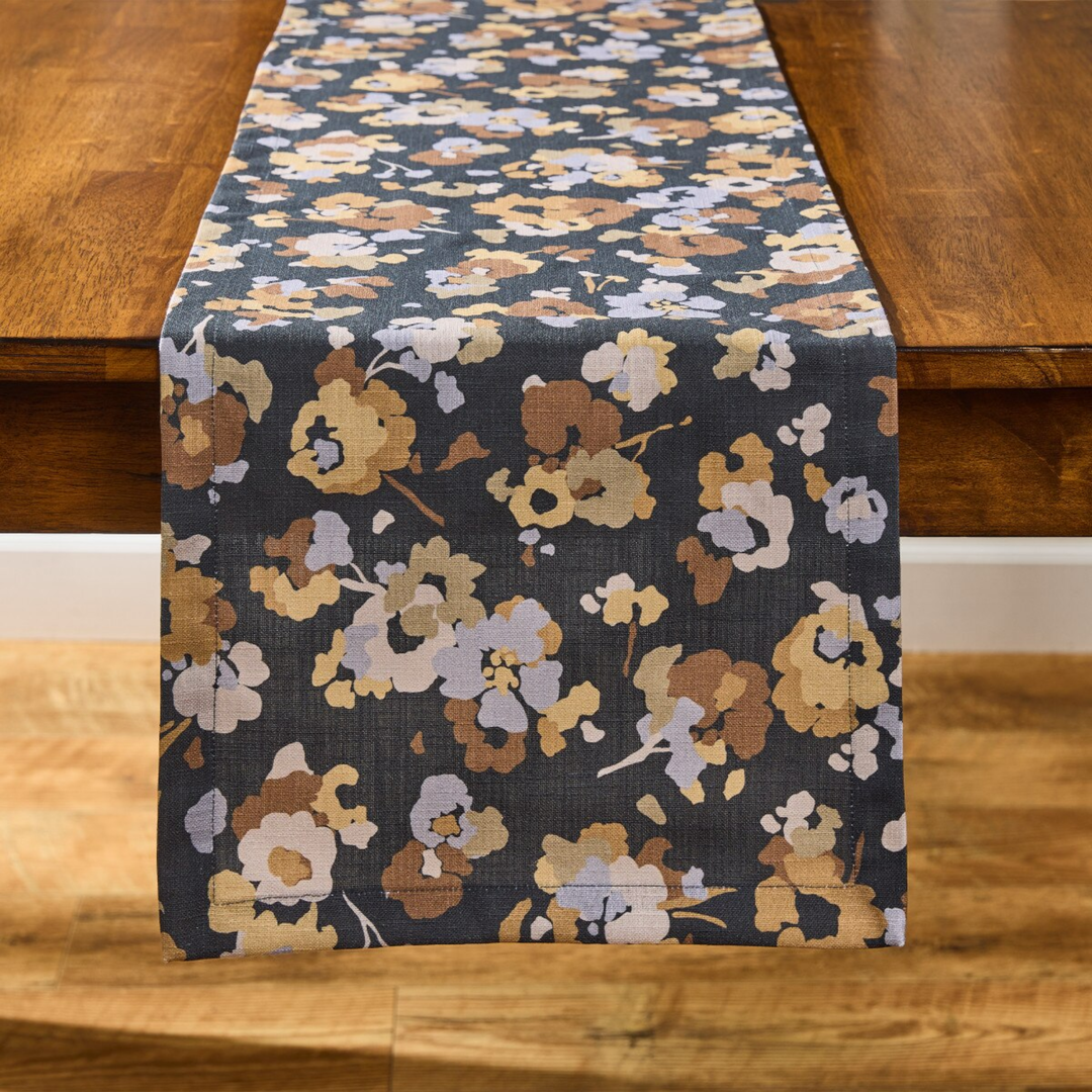 A floral table runner with a dark navy background and a pattern of beige, brown, and light lavender flowers, draped over a wooden dining table.