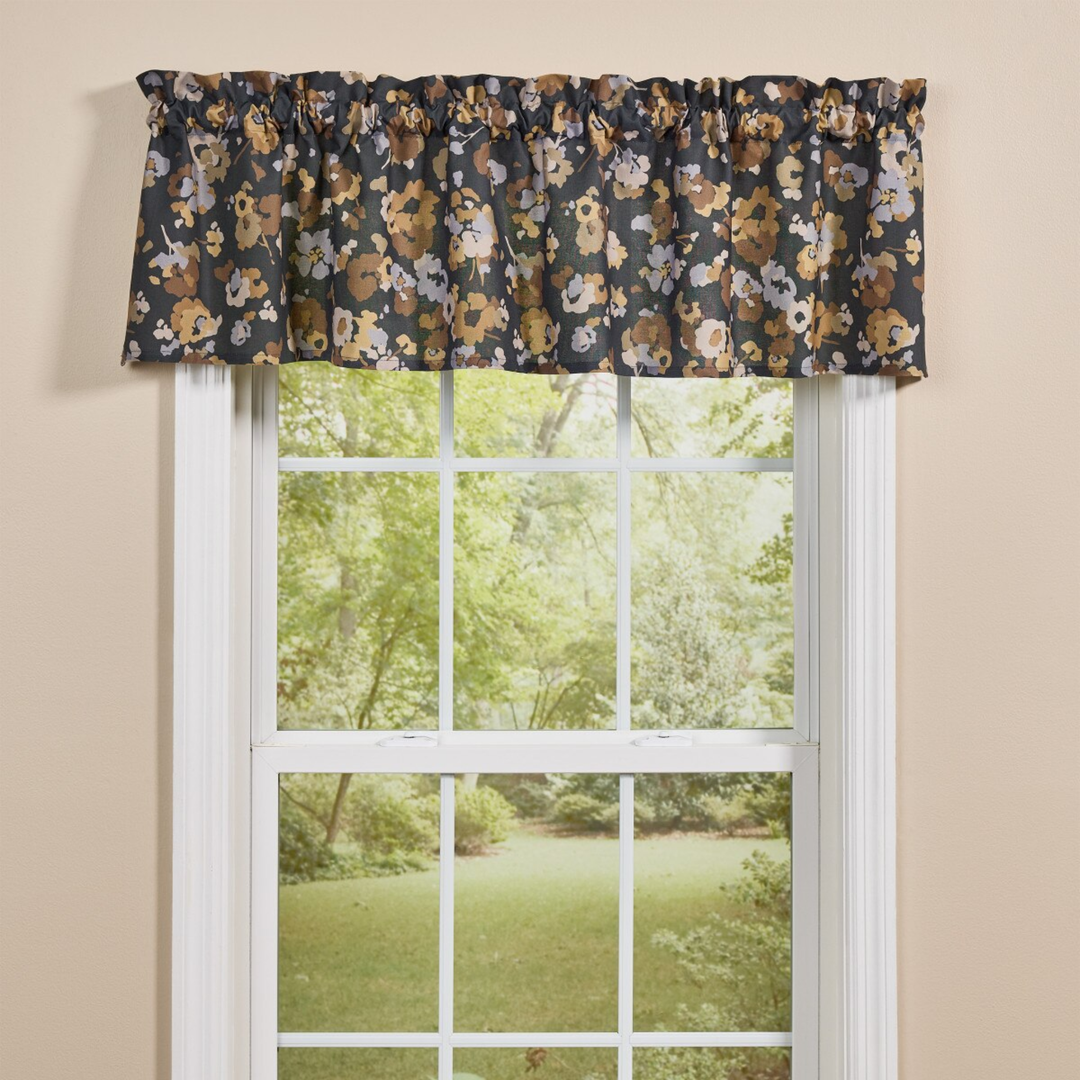 A floral valance curtain with a dark gray background and tan, beige, and white flowers, hung on a window with a view of a green garden outside.