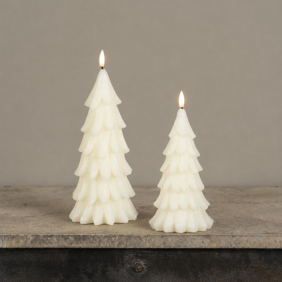 Two white Christmas tree-shaped candles with lit flames, standing on a rustic wooden surface against a neutral gray background.