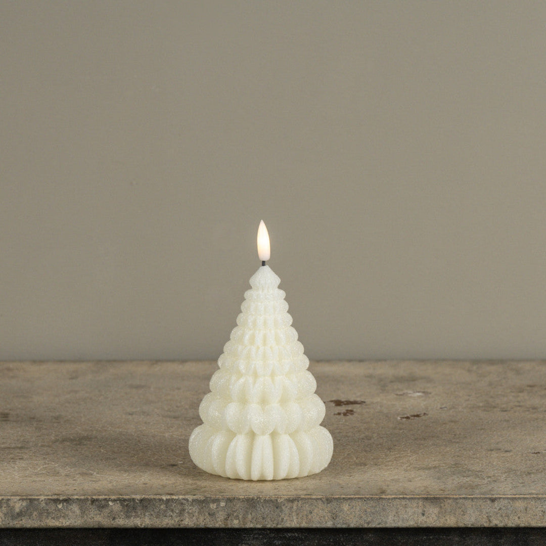 A lit white candle shaped like a small tree, standing on a rustic stone surface against a plain beige wall.