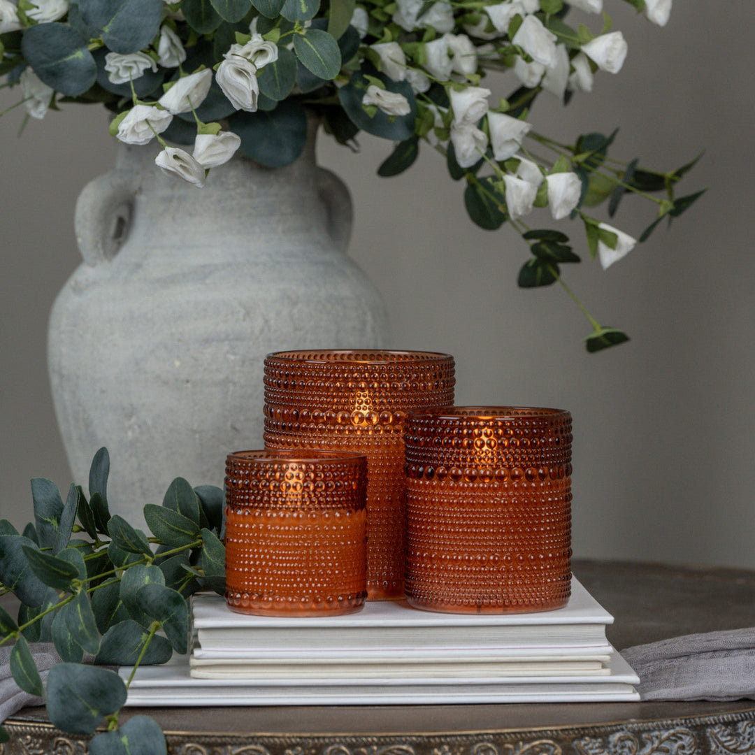 Set of three textured orange glass vases on a surface with a gray vase and greenery in the background.