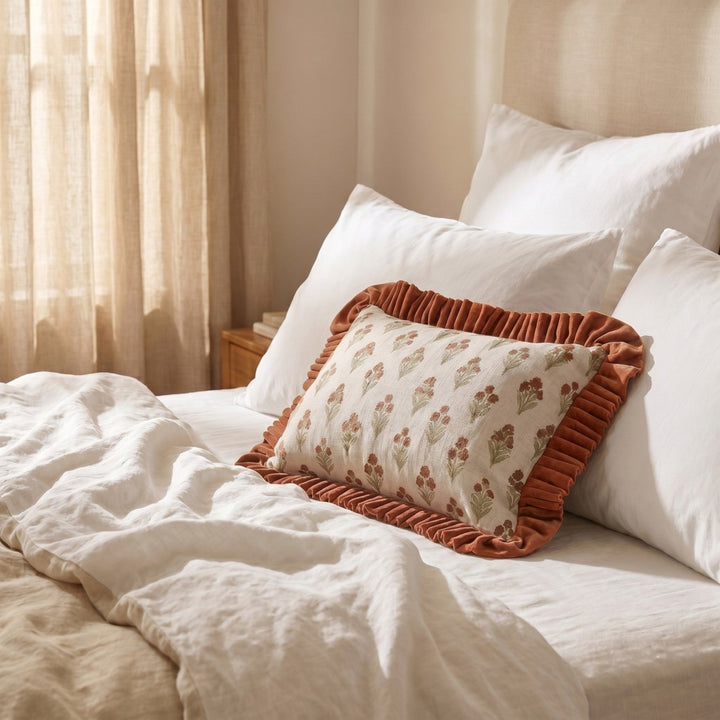 A decorative rectangular throw pillow with a cream floral pattern and rust-colored ruffled trim, styled on a bed with white and beige bedding in a softly lit bedroom.
