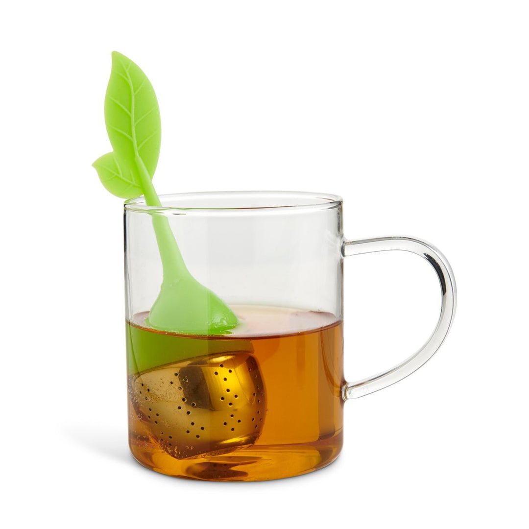 A clear glass mug with a handle, filled with amber tea and featuring a bright green silicone leaf-shaped tea infuser resting inside the mug, shown on a white background.