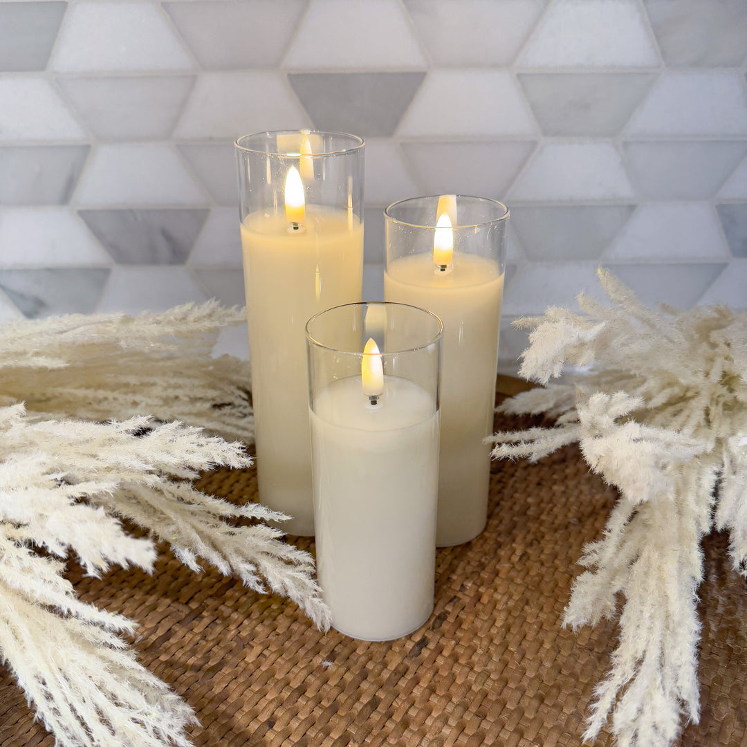 A set of three battery-operated candles arranged on a surface with decorative foliage, with each candle housed in a clear glass holder.
