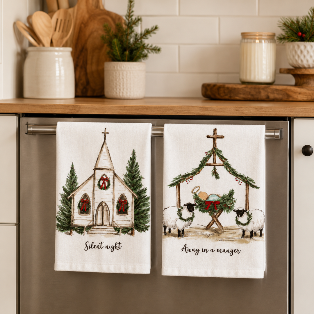 Two white kitchen towels hanging on a stainless steel dishwasher in a kitchen setting. The left towel features an illustration of a small church with Christmas wreaths and the text 'Silent night'.
