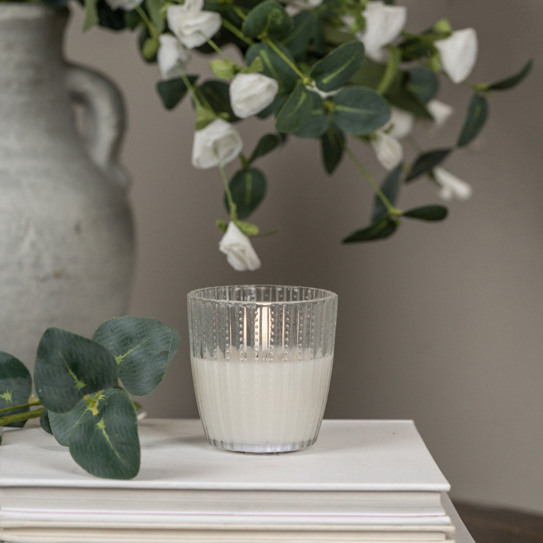 Clear glass candle holder with white candle on a surface, with flowers and a vase in the background.