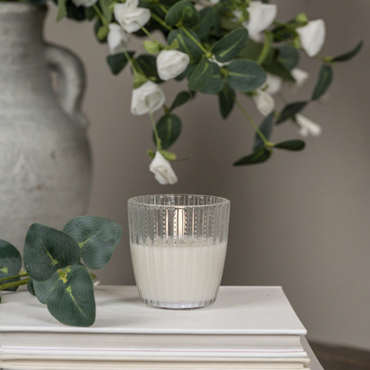 Clear glass candle holder with white candle on a surface, with flowers and a vase in the background.