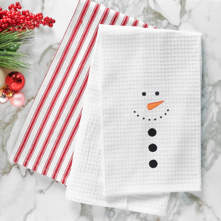A folded white waffle-weave kitchen towel with an embroidered snowman face, styled on a white marble surface with holiday greenery, red berries, candles, and a red-and-white striped cloth.