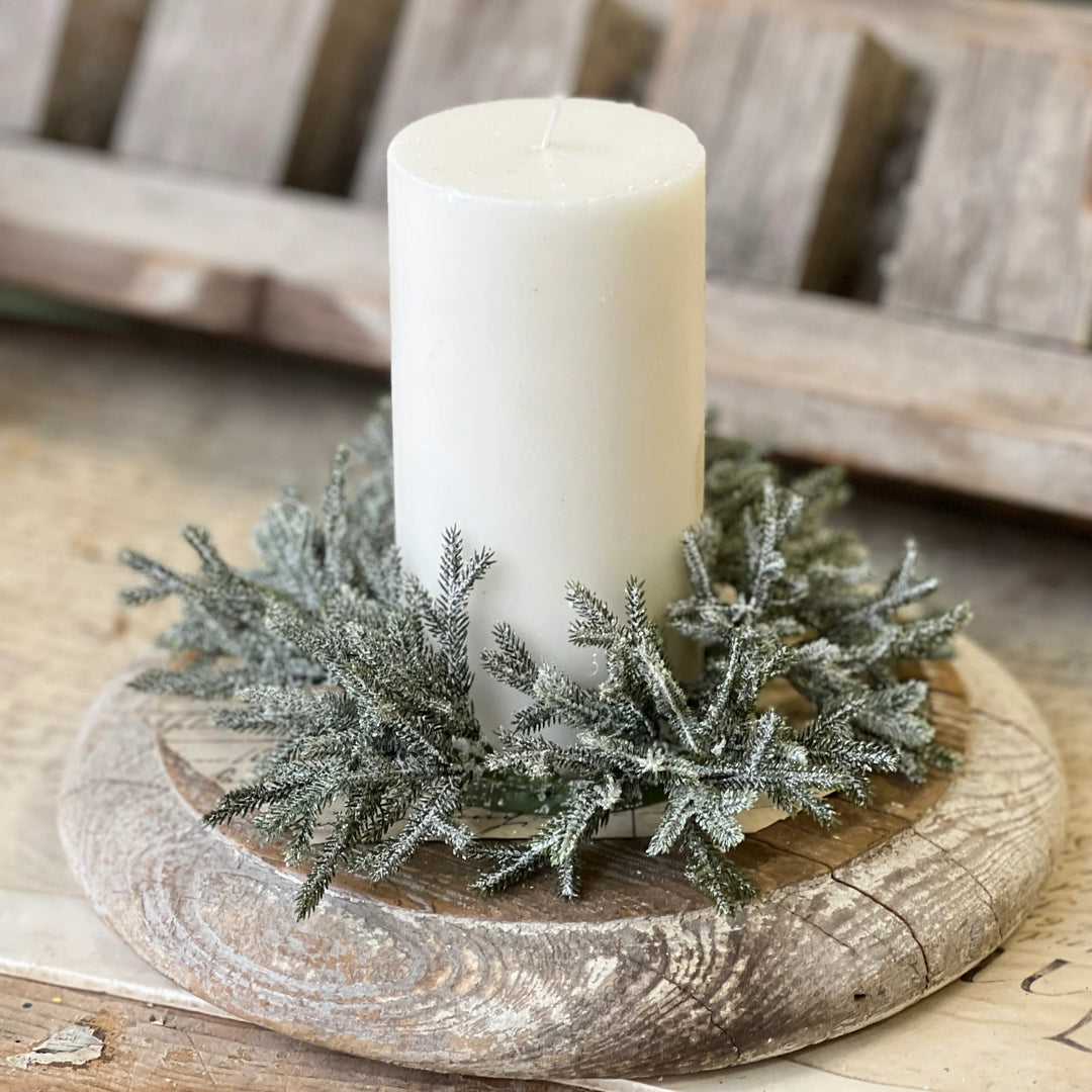 A white pillar candle displayed in a rustic wooden holder with frosted green pine branches, set on a weathered wooden surface.