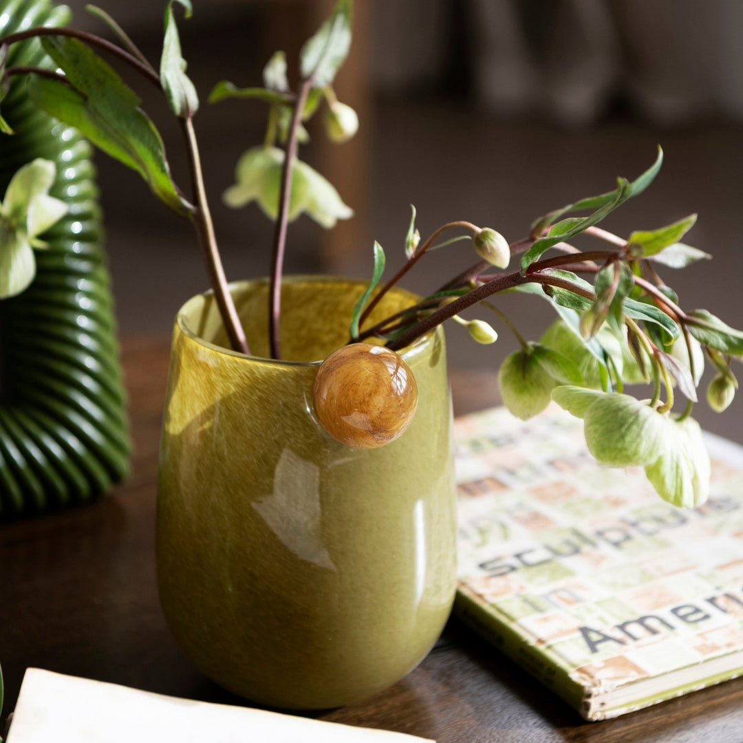An olive green ceramic bud vase with a glossy finish, holding delicate green and white spring flowers, set on a dark wooden table next to a book.